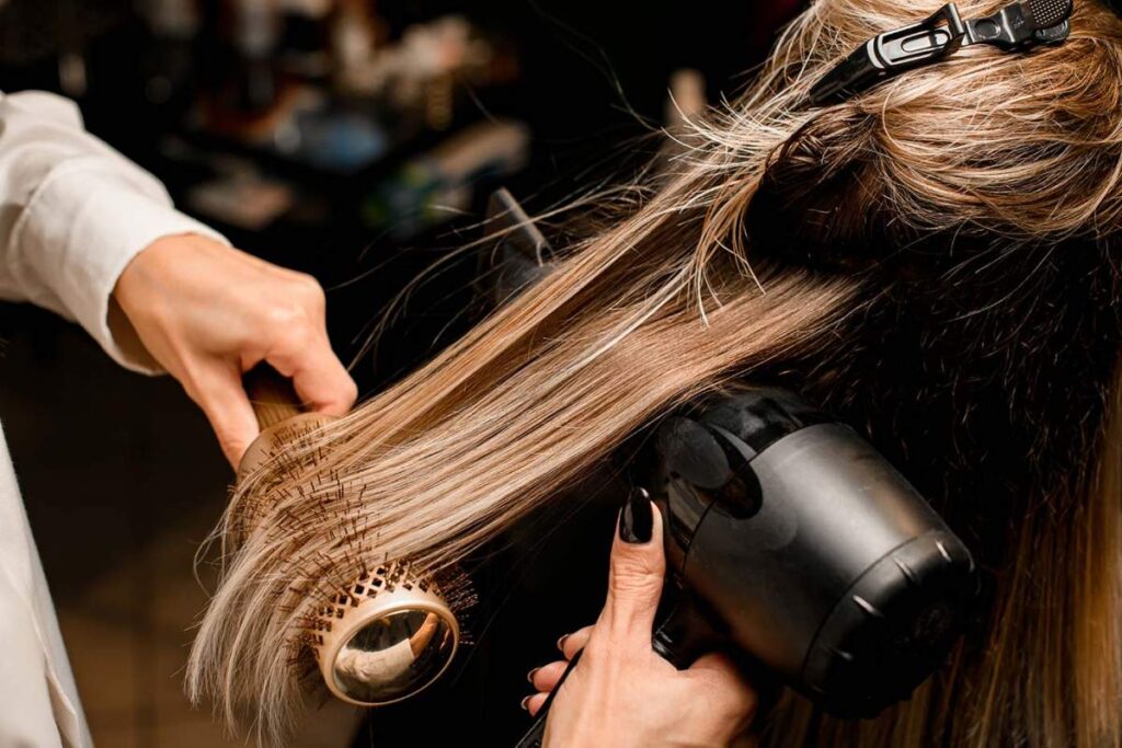 Person lifting fine hair at the roots with a round brush while blow-drying in a bright bathroom.