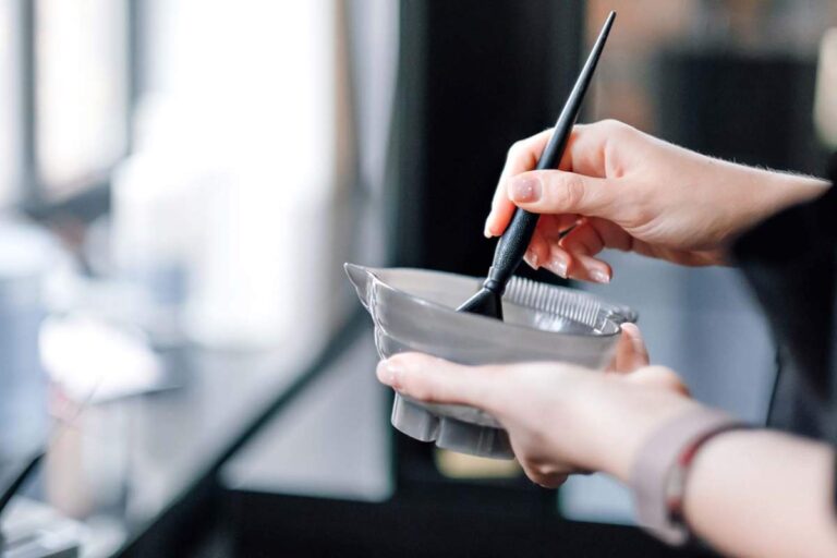 Person applying a creamy hair mask to damp hair in a bright bathroom with a wide-tooth comb nearby.